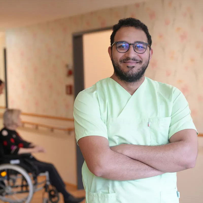 Pflegehelfer bei PFLEGEN & WOHNEN HAMBURG unterstützt Bewohnerin im Rollstuhl in Pflege, Betreuung und Wohlbefinden.